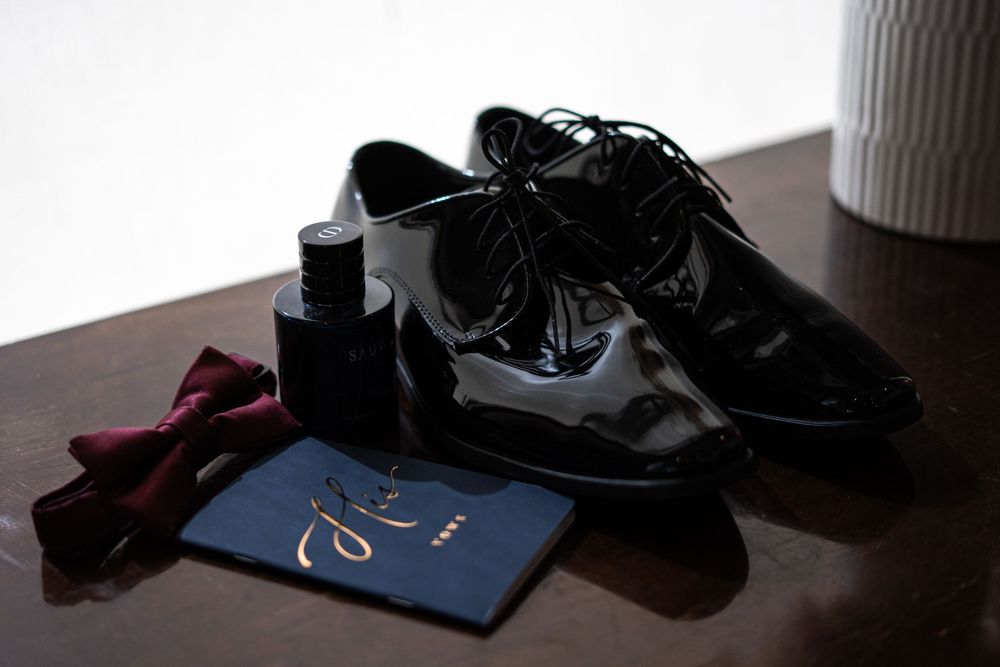 Shiny black patent leather oxford shoes with red bow tie, cologne bottle, and navy invitation on wooden surface.