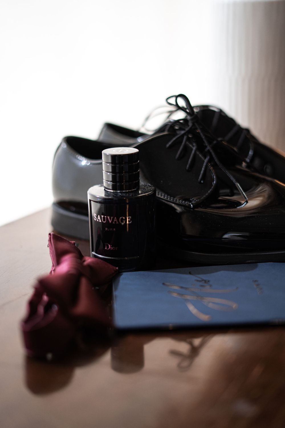 Dior Sauvage cologne beside black patent leather dress shoes and burgundy tie on a table.