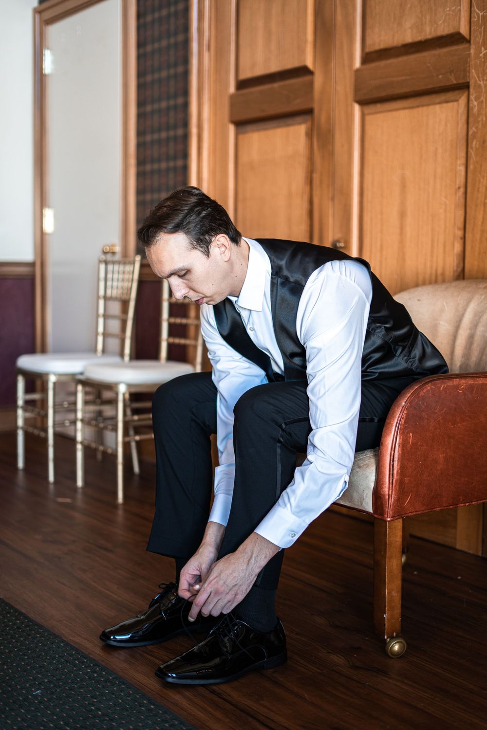 A groom in a black vest and tie sits on a leather bench, tying his formal black dress shoes before his wedding.
