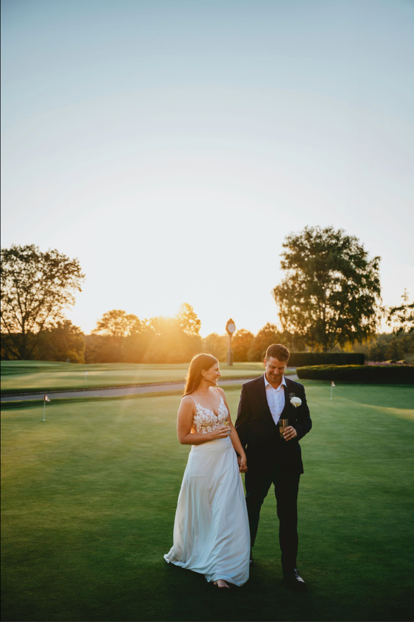 Bride and groom walking on golf course at golden hour sunset, she in white gown, he in black tuxedo.
