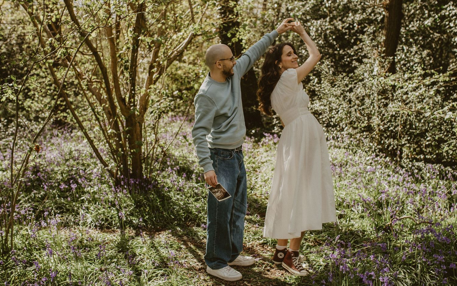 Couple dancing together in a sunlit woodland garden, man in blue jeans spinning woman in white dress among wildflowers.