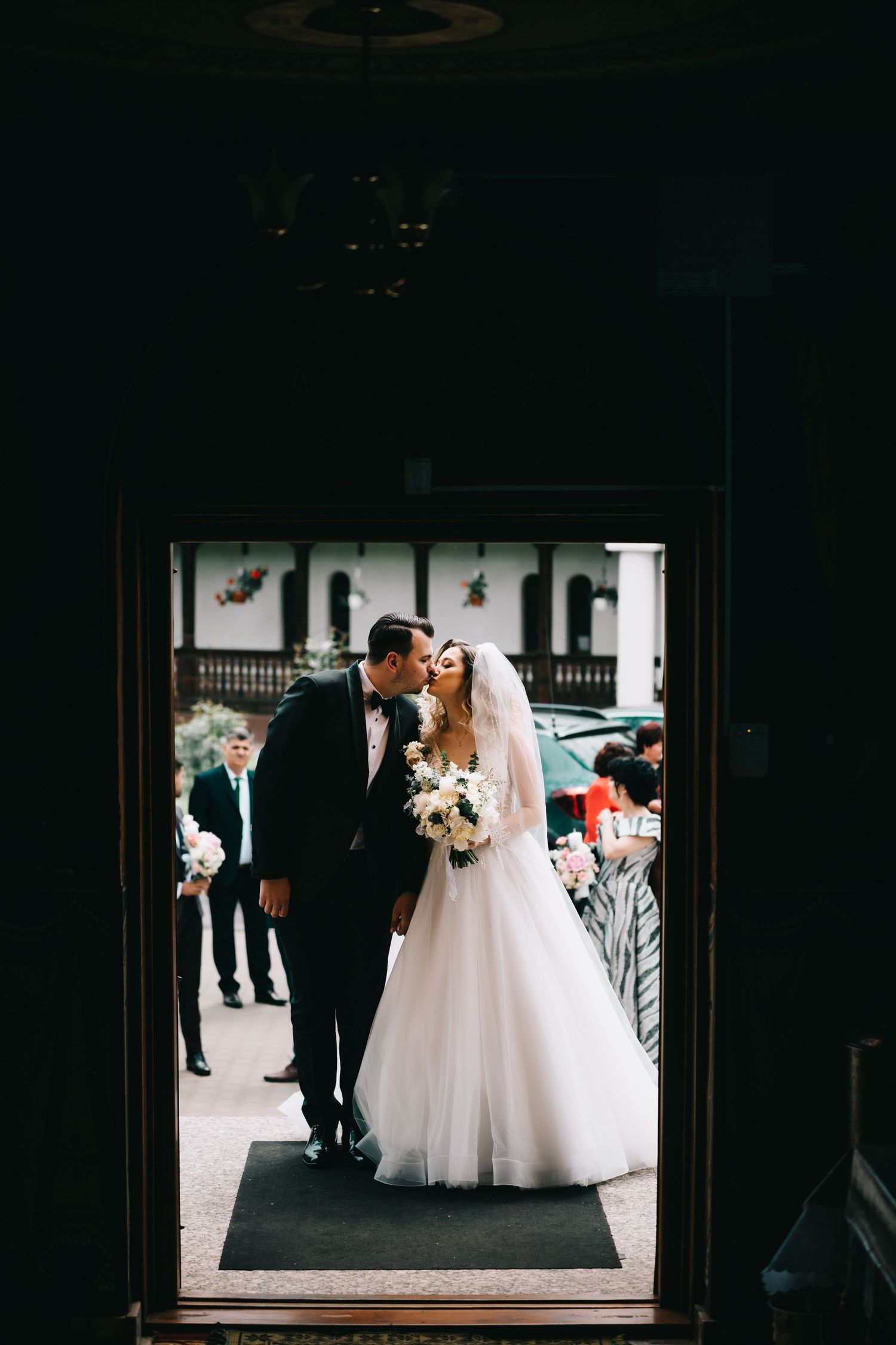 Elegant bride and groom pose together against white paneled wall, bride holding white floral bouquet.
