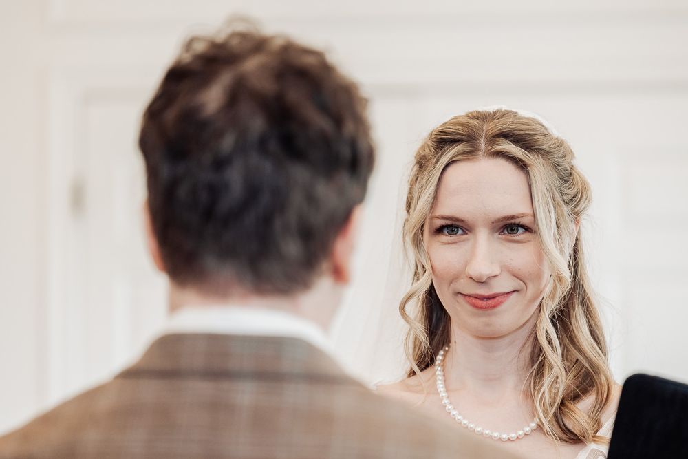 Bride smiling warmly at groom during wedding ceremony, wearing pearl necklace with elegant half-up hairstyle.