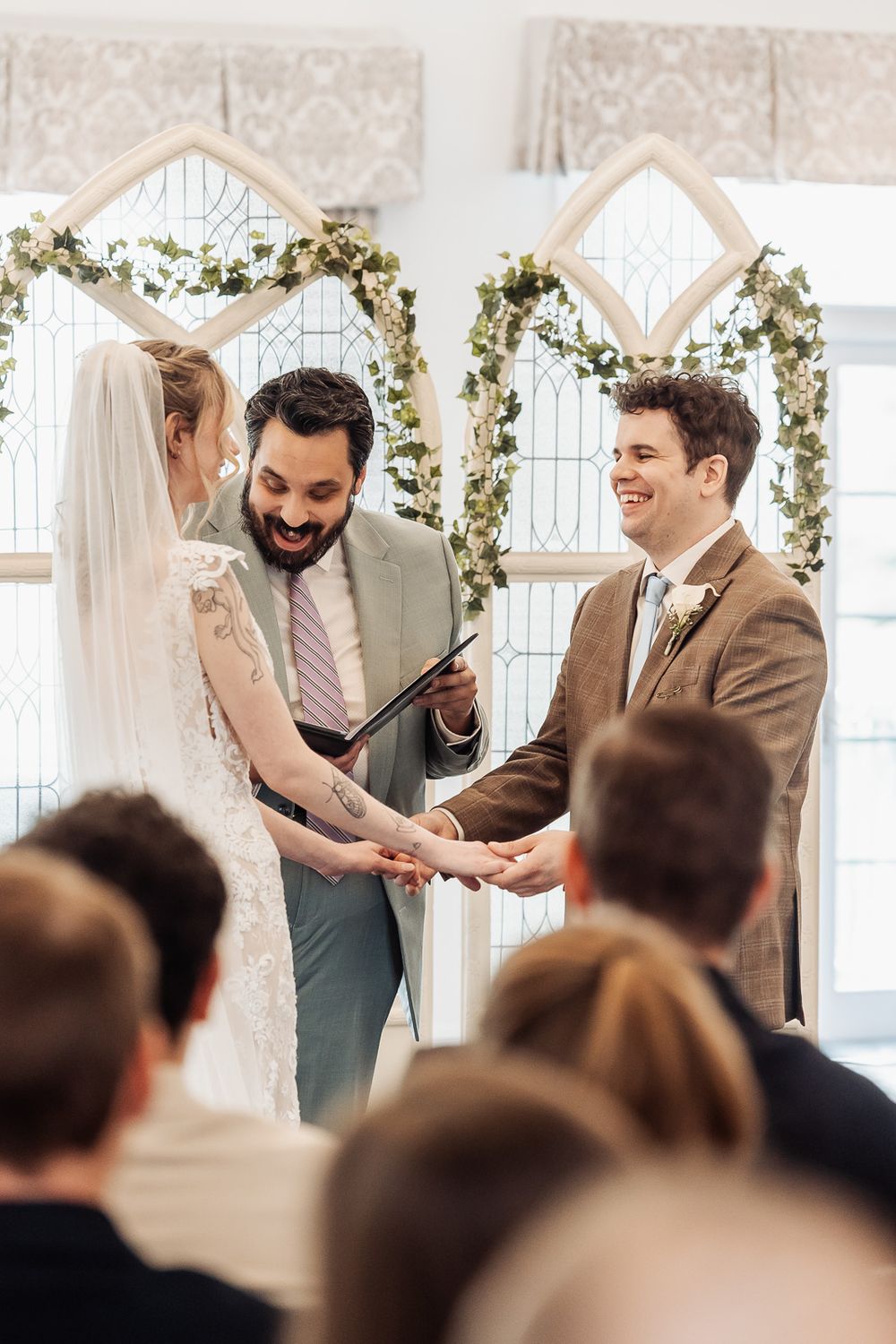 Smiling groom holds bride's hands at altar during indoor wedding ceremony with floral arch backdrop.