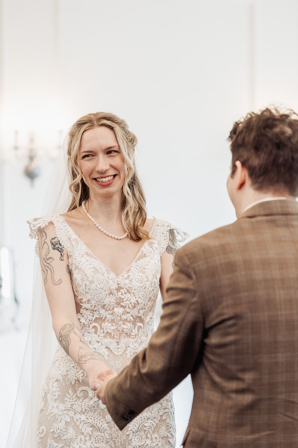 Smiling bride in lace wedding dress with tattoos holds hands with groom in brown plaid suit during wedding ceremony.