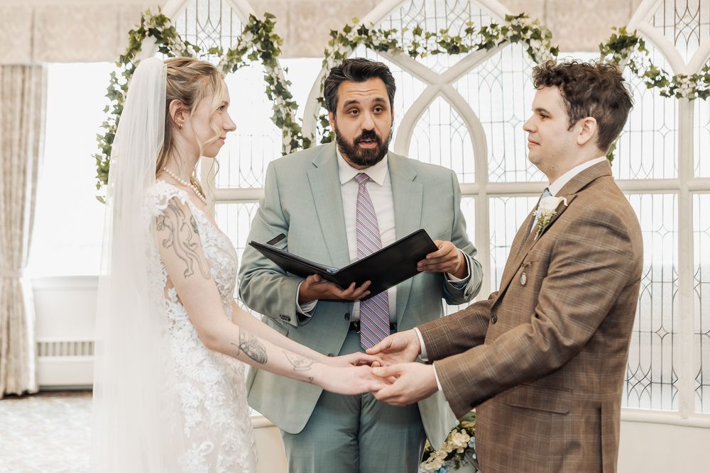 Bride and groom exchange vows at elegant indoor wedding ceremony with officiant holding book before floral arch.