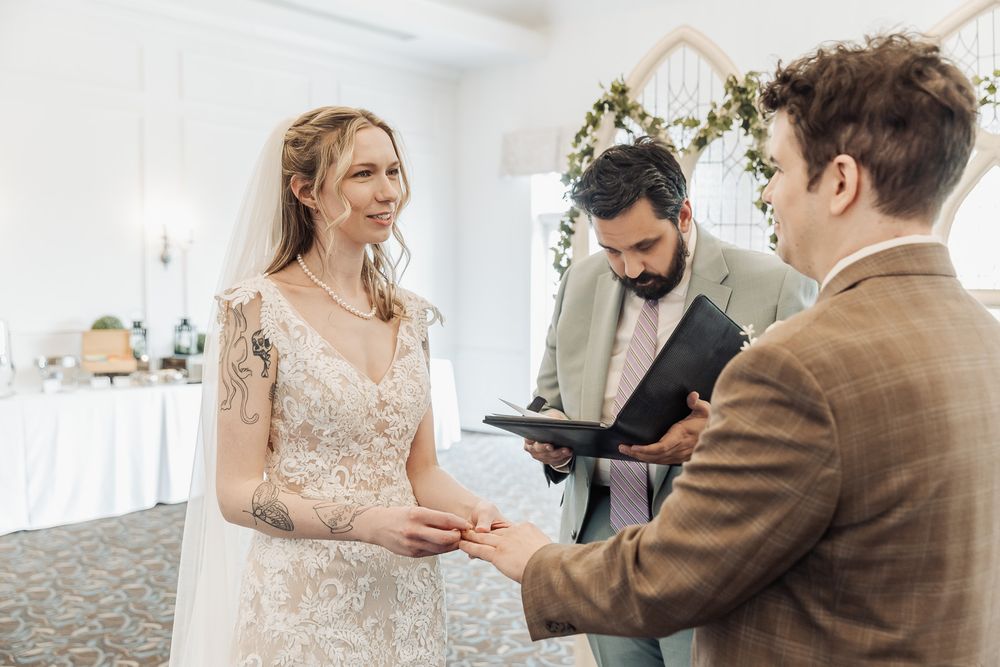 Bride and groom exchanging rings during wedding ceremony with officiant holding book indoors.