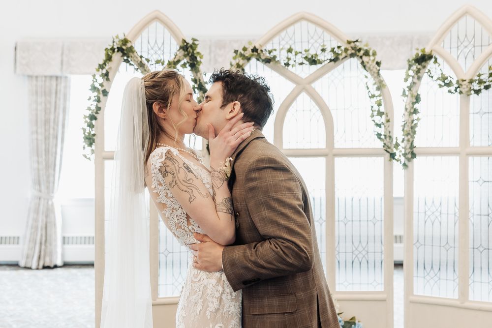 Bride and groom share first kiss at wedding altar decorated with white arch and green floral garland.