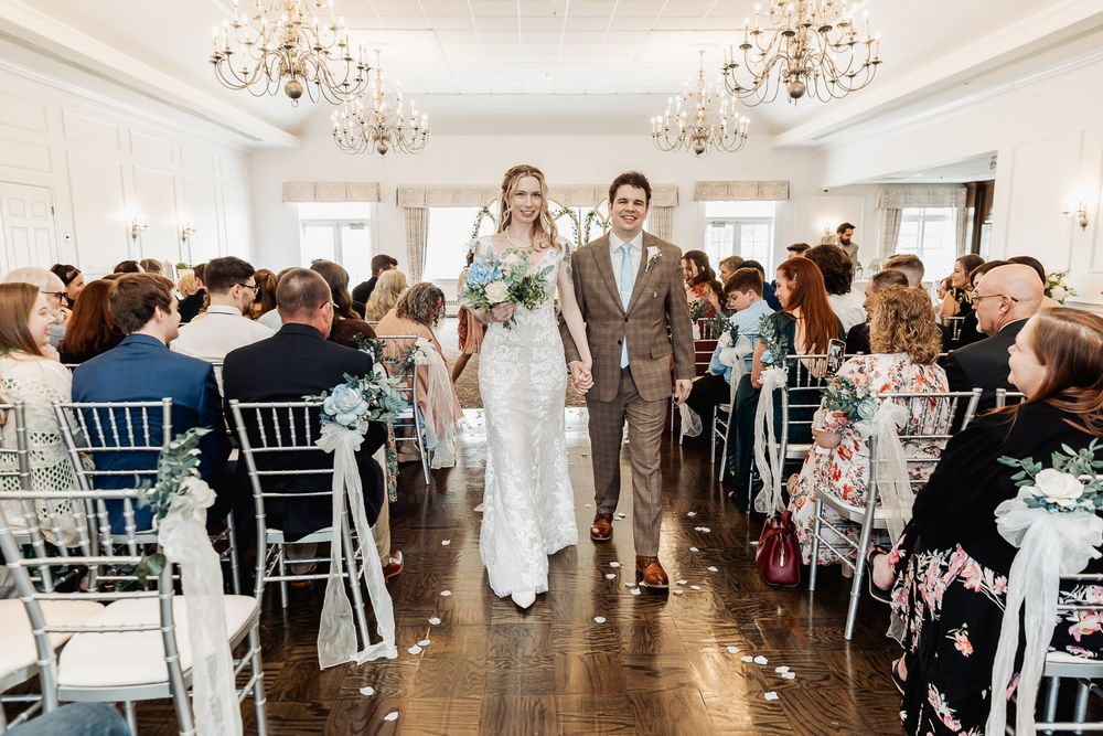 Bride and groom walk down the aisle at an elegant indoor wedding ceremony with chandeliers and seated guests.