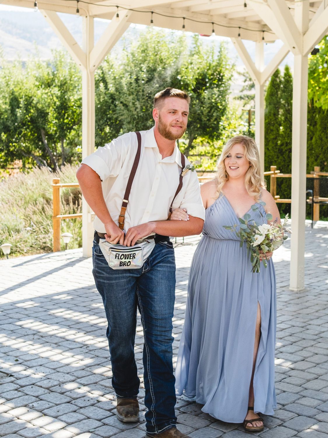 Two smiling groomsmen in white shirts and jeans pose outdoors, one wearing suspenders and a Flower Bro fanny pack.