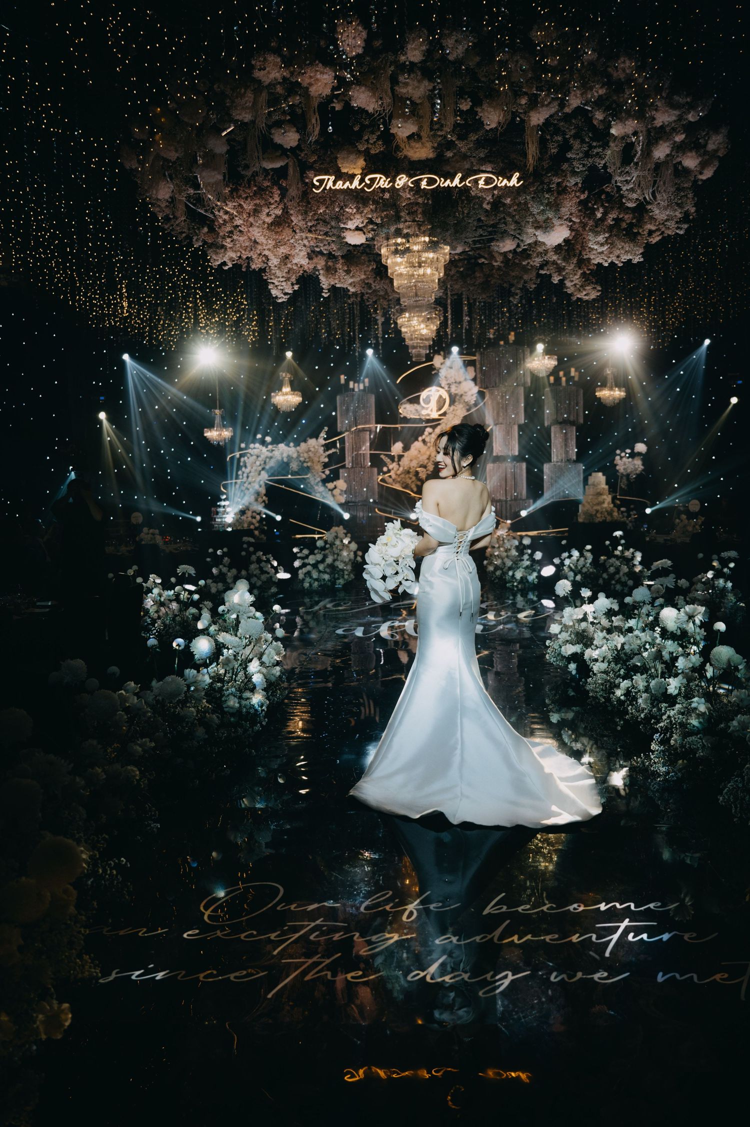 Bride in white mermaid gown standing in dramatic floral wedding venue with chandeliers and stage lighting.