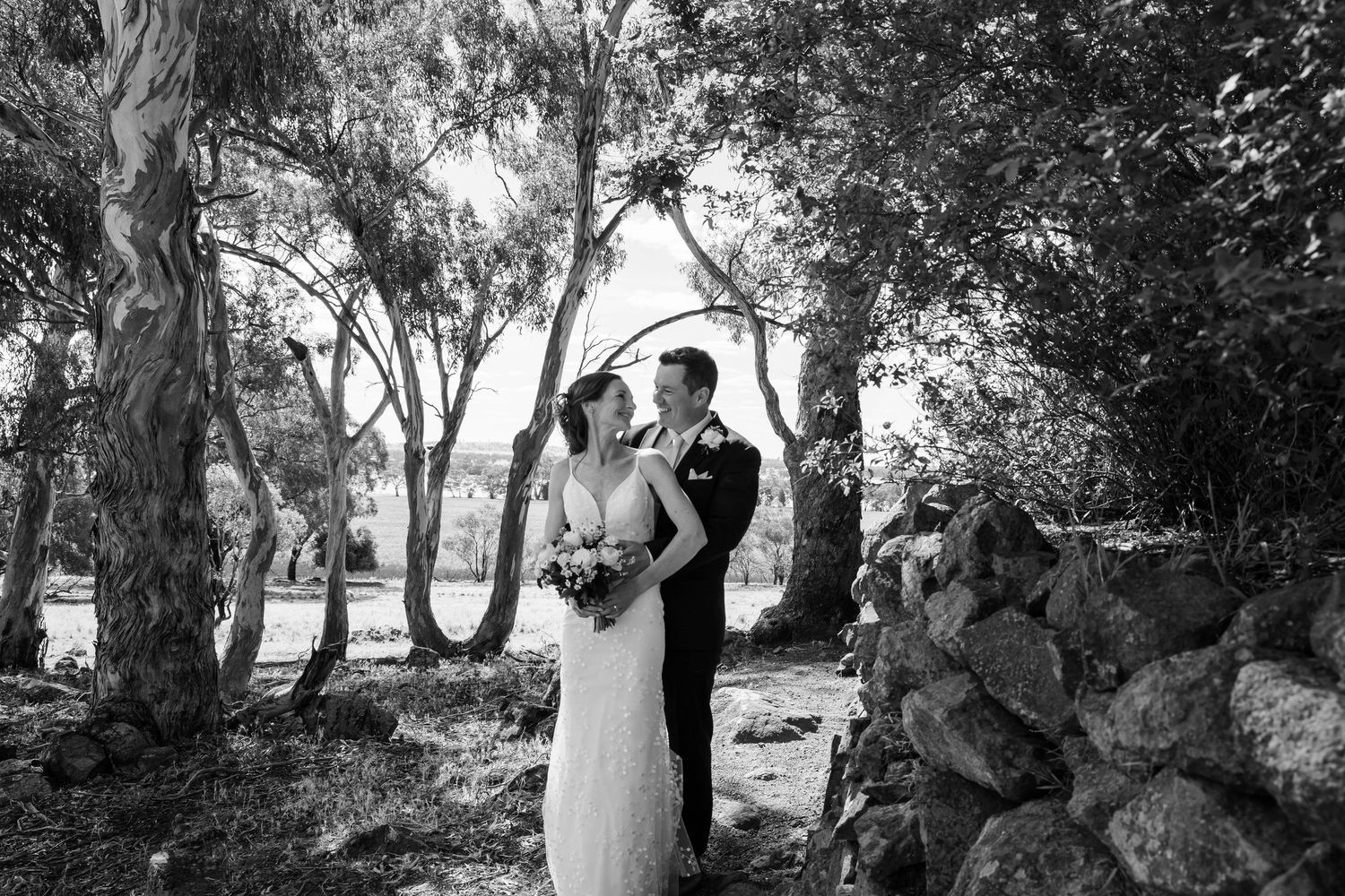 A bride and groom share a romantic kiss in a lush garden beside a charming white cottage adorned with flowers.