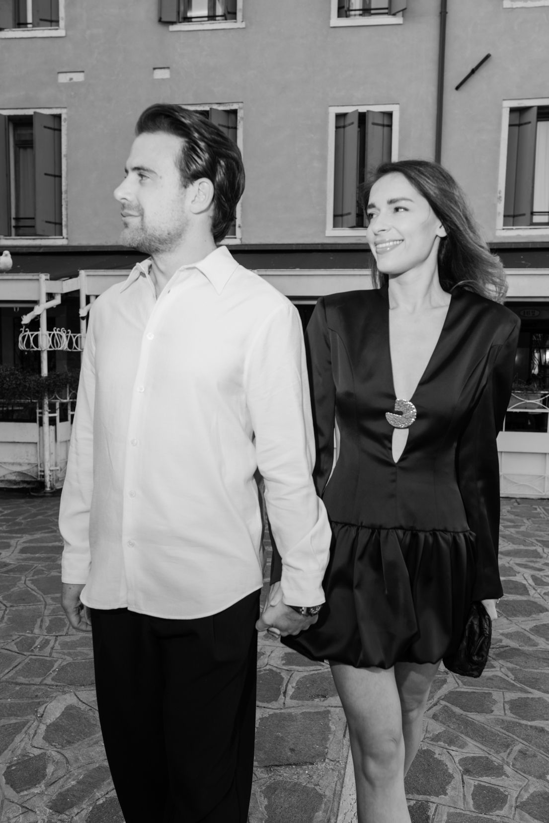 Black and white photo of an elegant couple holding hands in front of a European building.