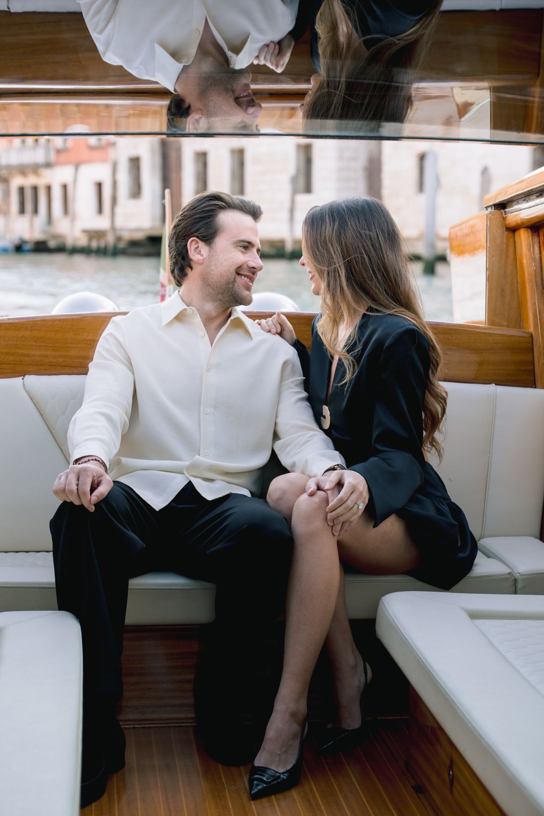 Elegant couple holding hands and smiling on a luxury boat in Venice, Italy, with canal views in the background.