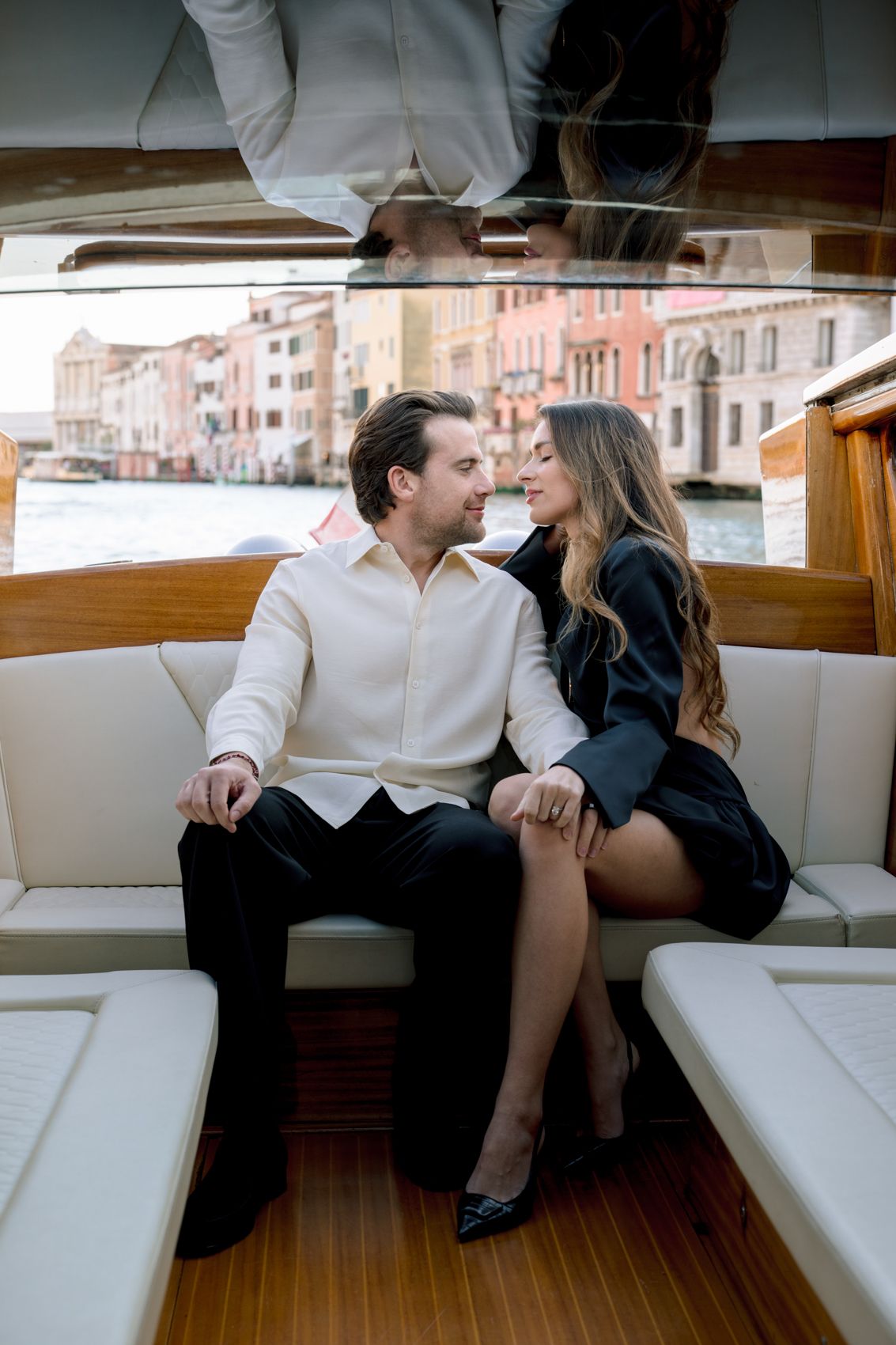 Couple sitting close together on a luxury boat in Venice canal, smiling at each other, man in white shirt, woman in black dress.