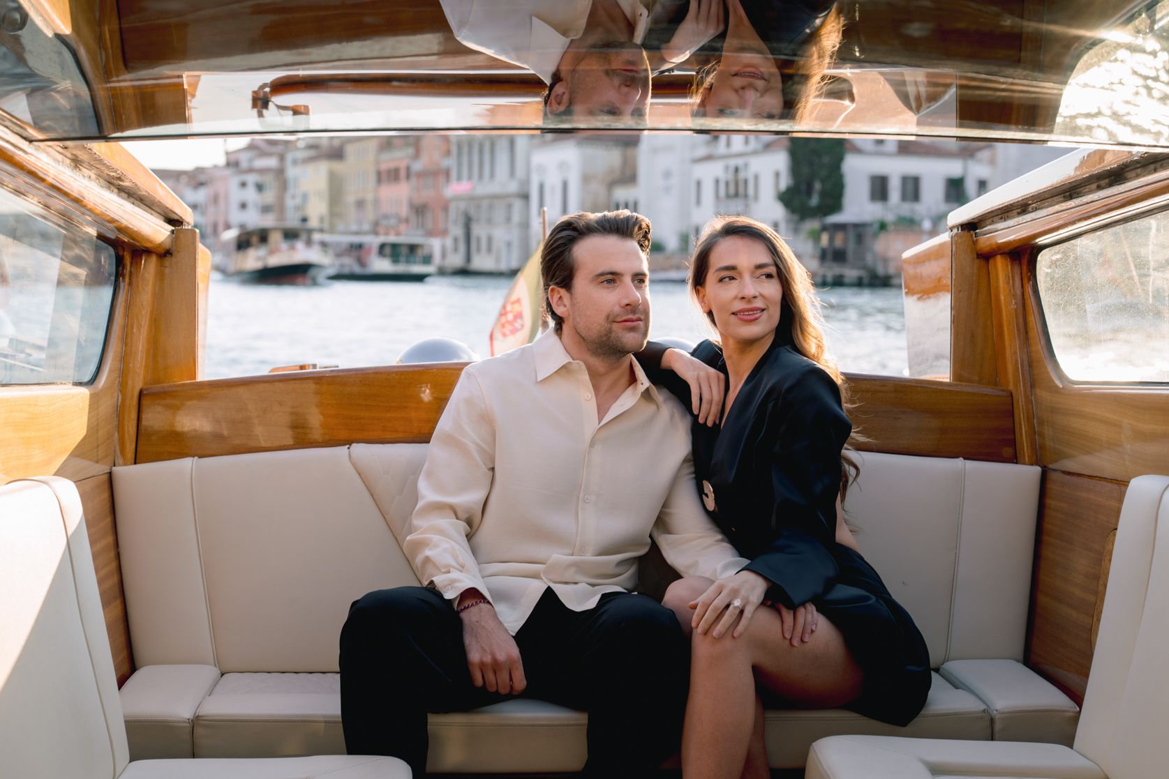 Couple sitting together on a luxury wooden boat in Venice canal, woman in black dress, man in cream shirt.