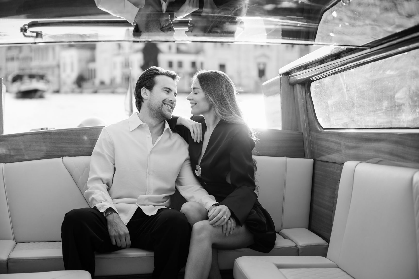 Black and white photo of a romantic couple sitting close together smiling inside a classic wooden boat.