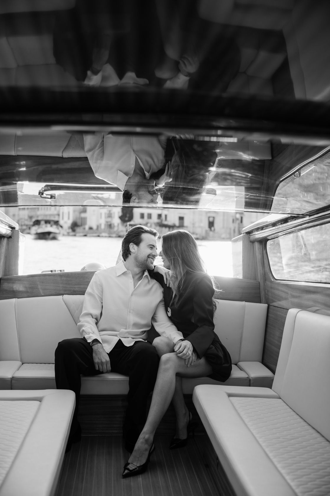 Black and white photo of a romantic couple sitting close together on a luxury boat, smiling and gazing at each other.