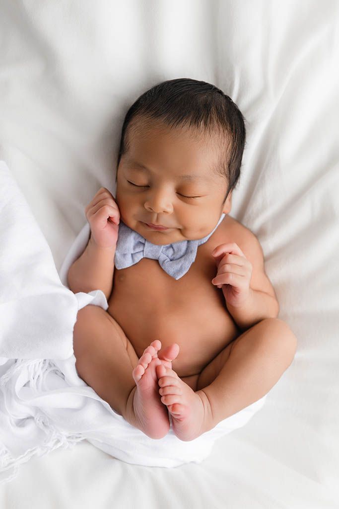 Sleeping newborn baby boy wearing a light blue bow tie, lying on a white blanket by Murrieta California photographer.