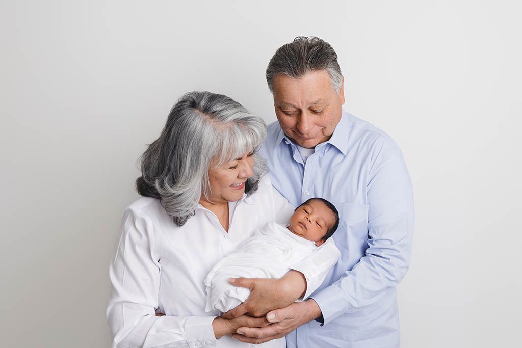 Grandparents lovingly holding a sleeping newborn baby wrapped in white against a light background by Murrieta CA newborn photographer.