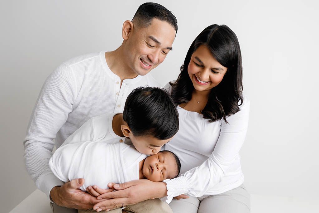 Family portrait with parents and son holding newborn baby boy, dressed in white, on white background by Murrieta newborn photographer.