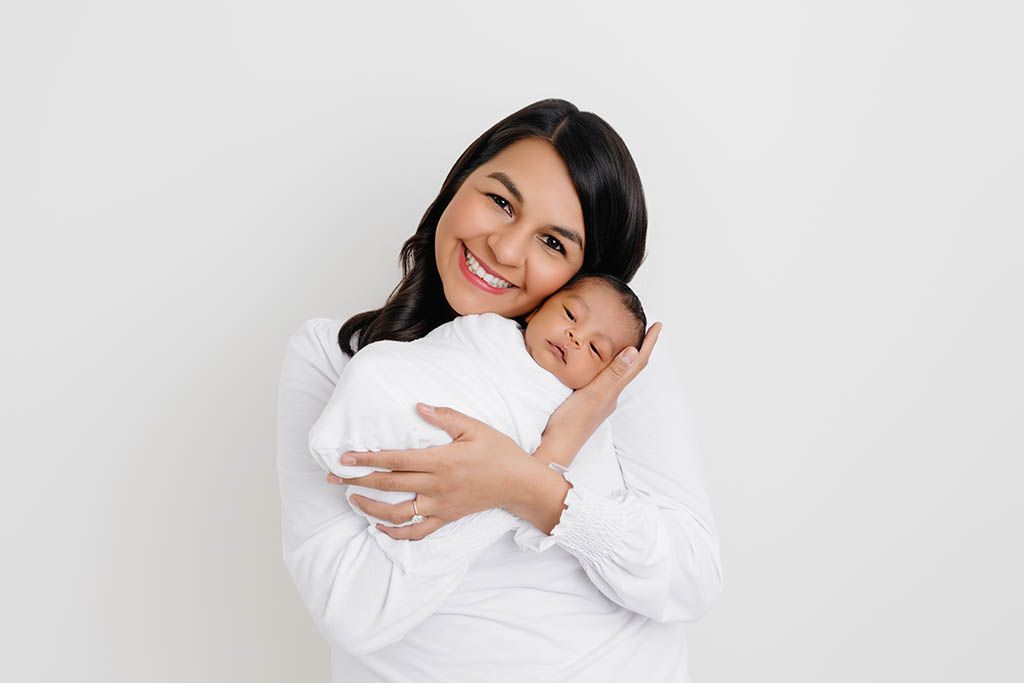 Smiling mom lovingly cradles sleeping newborn baby boy against white background by professional Murrieta newborn photographer.