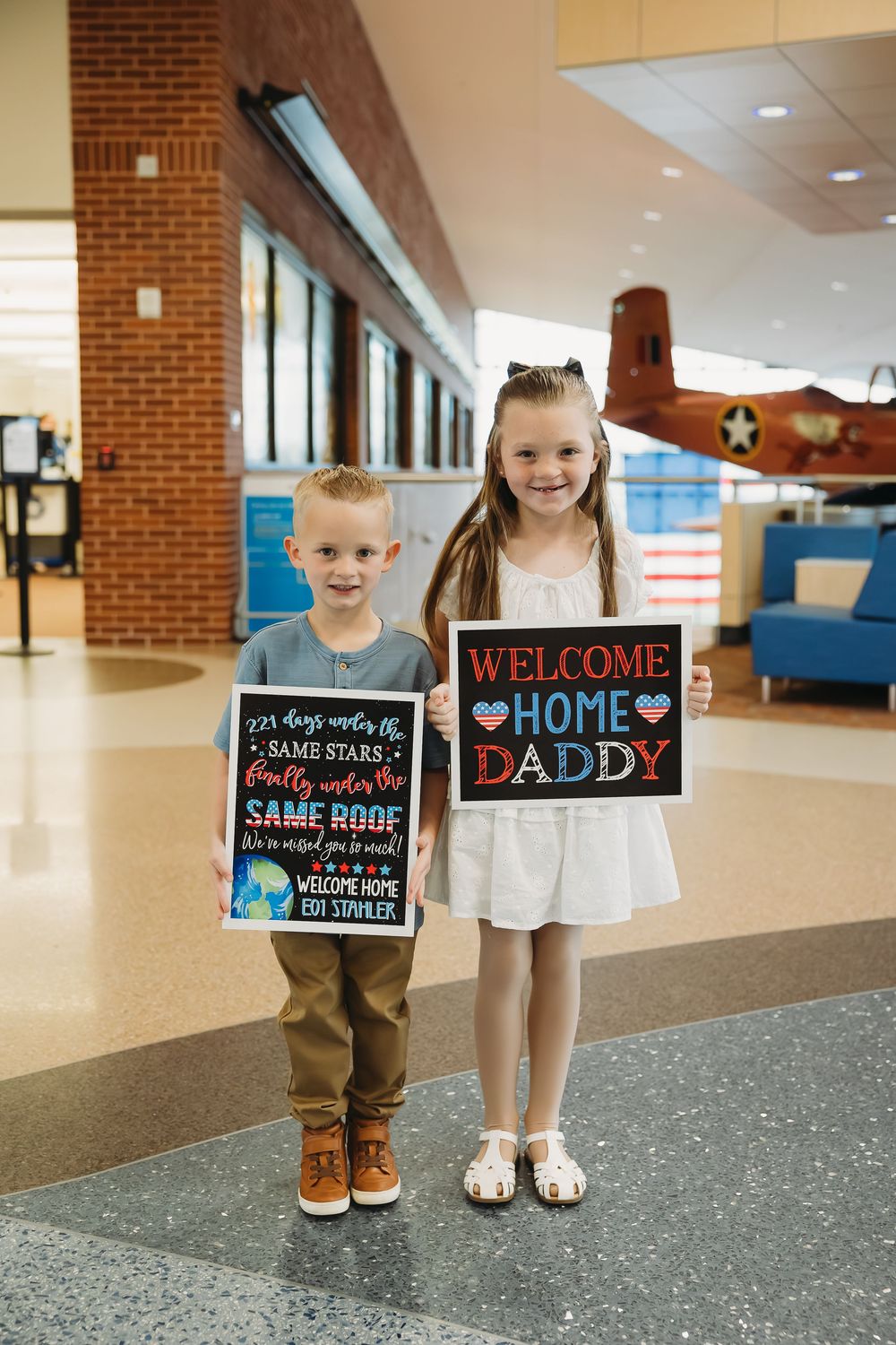 Two children holding Welcome Home Daddy signs in a military building lobby, awaiting a parent's homecoming.