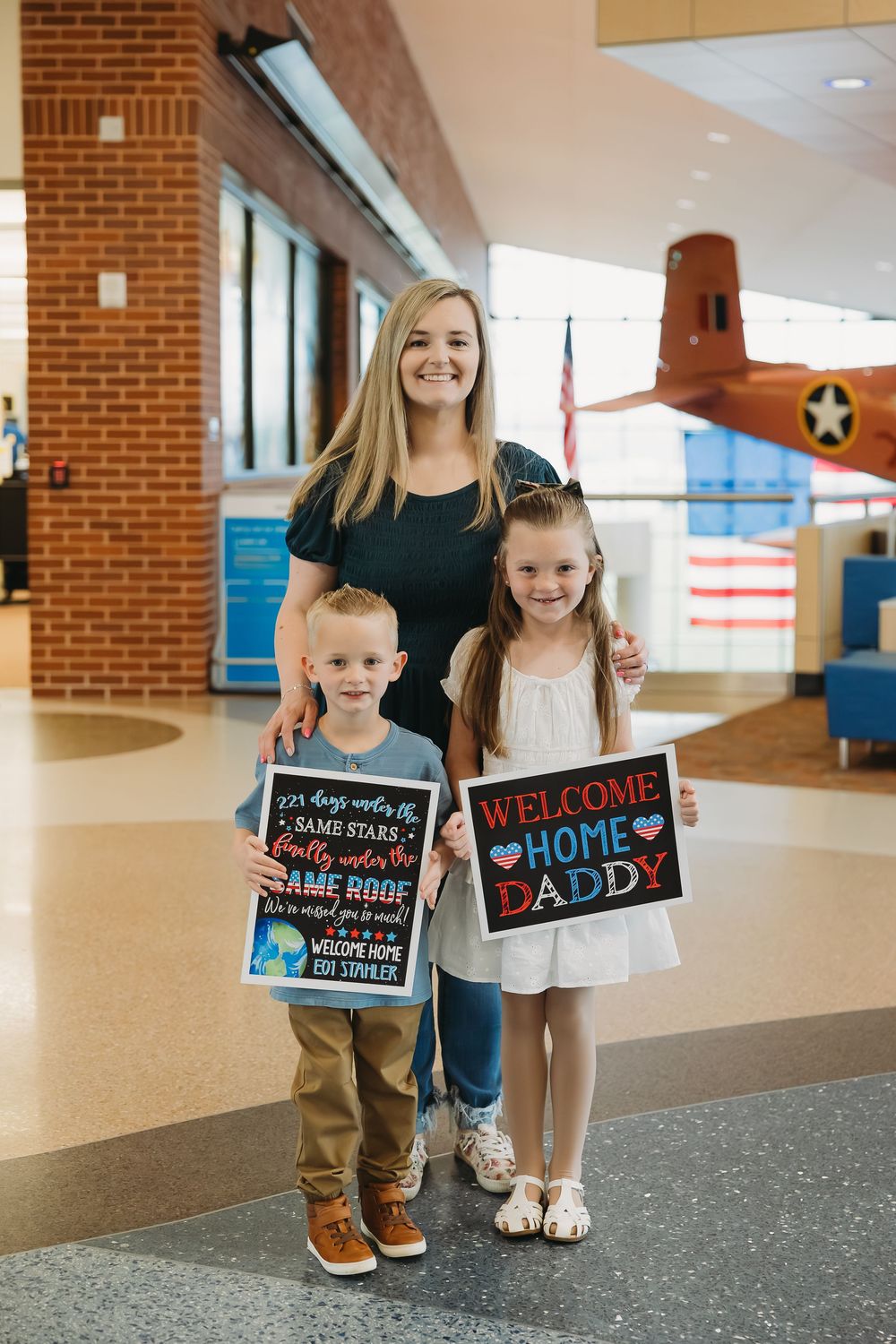 A woman and two children hold Welcome Home Daddy signs at a military airport homecoming reunion.