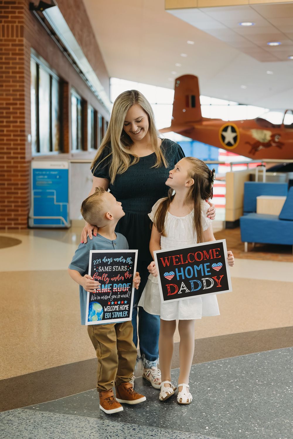 A mother and two children hold welcome home daddy signs in an airport, smiling during a military homecoming.