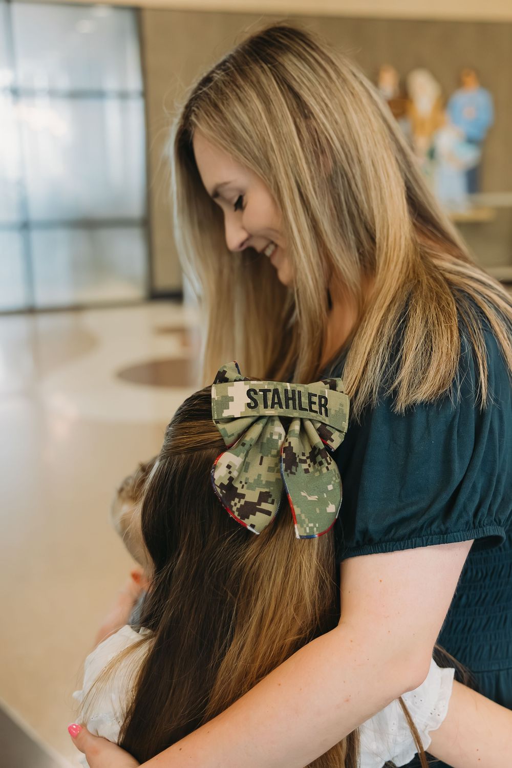 Woman smiling while hugging a child, wearing a camouflage bow hair clip with the name STAHLER on it.