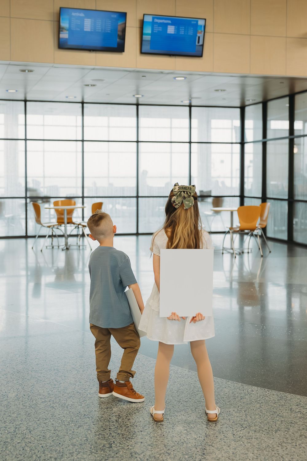 Two children holding hands in an airport terminal, girl in white dress holding a blank sign.