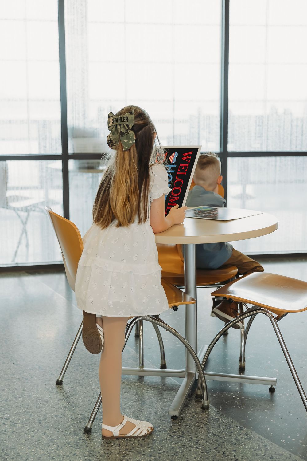 Two children sit at a round table in a bright room, with a girl in a white dress reading a colorful book.