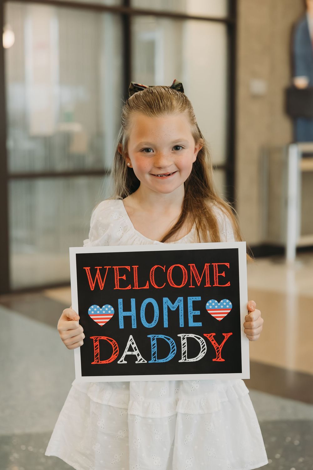Young girl in white dress smiling while holding a Welcome Home Daddy sign with American flag hearts.
