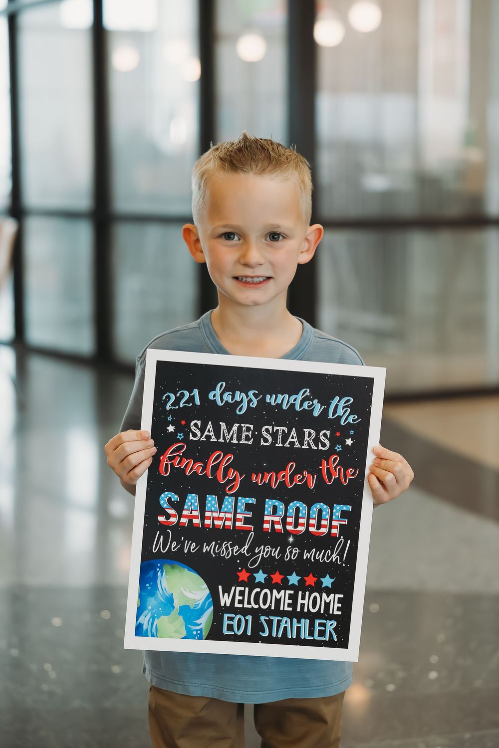 Young boy holding a Welcome Home military sign reading 221 Days Under the Same Stars, Finally Under the Same Roof.