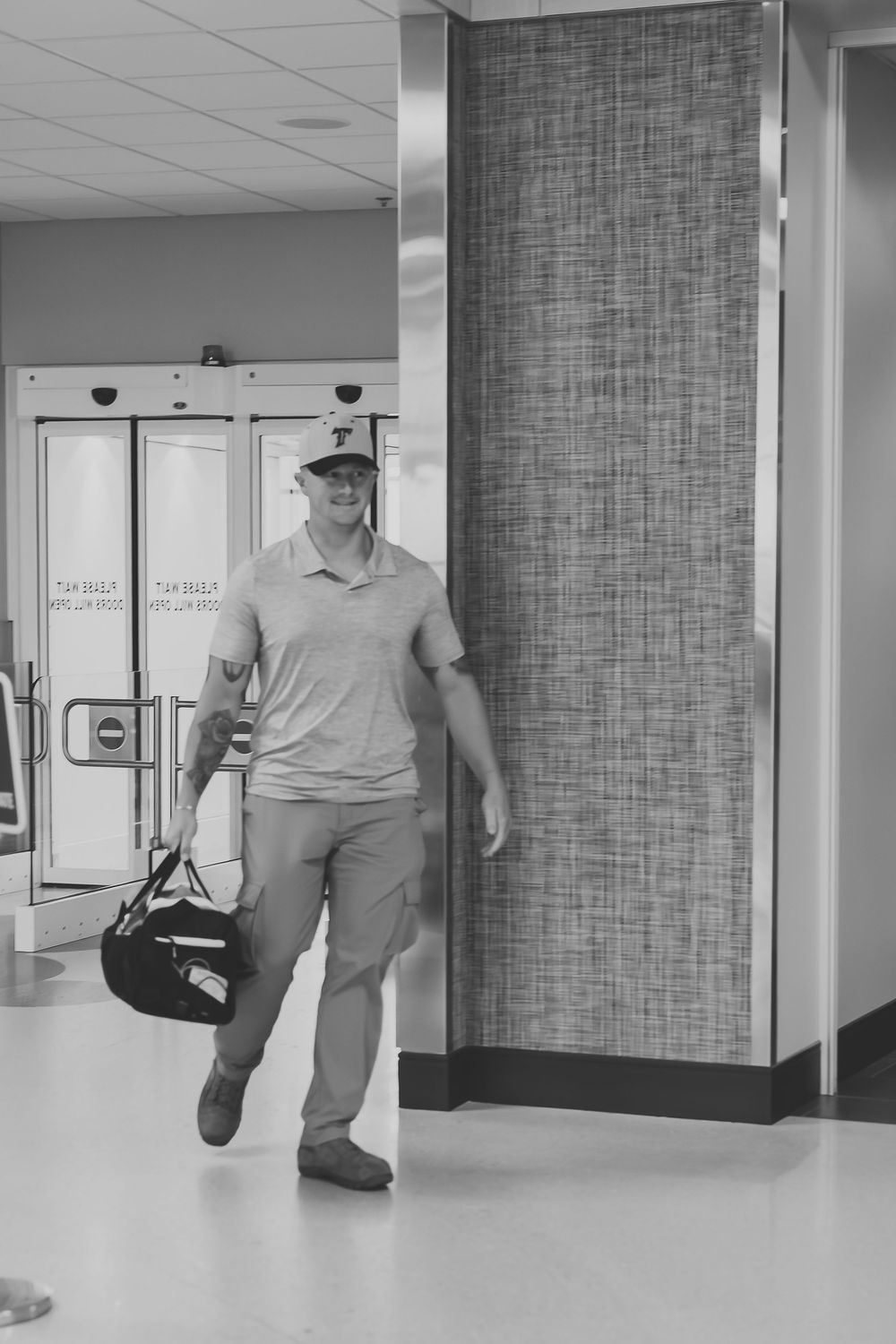 Black and white photo of a smiling man in a cap carrying a duffel bag walking through a modern building entrance.