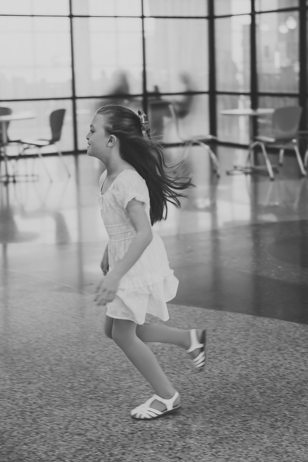 Black and white photo of a young girl in a white dress running through a modern building interior.