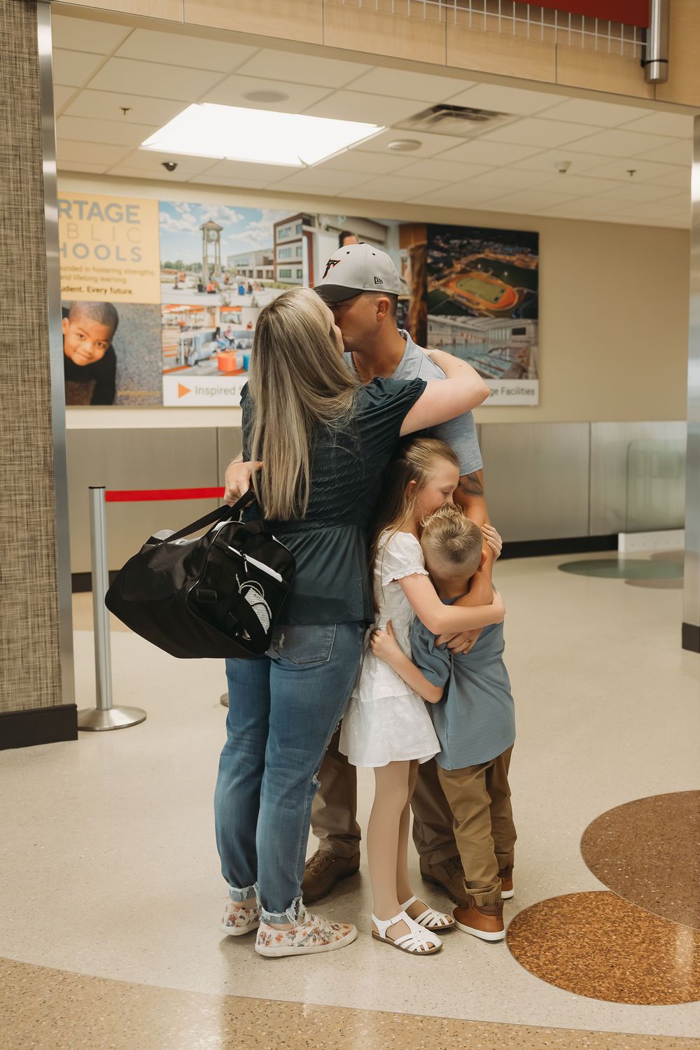 Family reunion hug at airport terminal, parents embracing two young children in emotional homecoming moment.