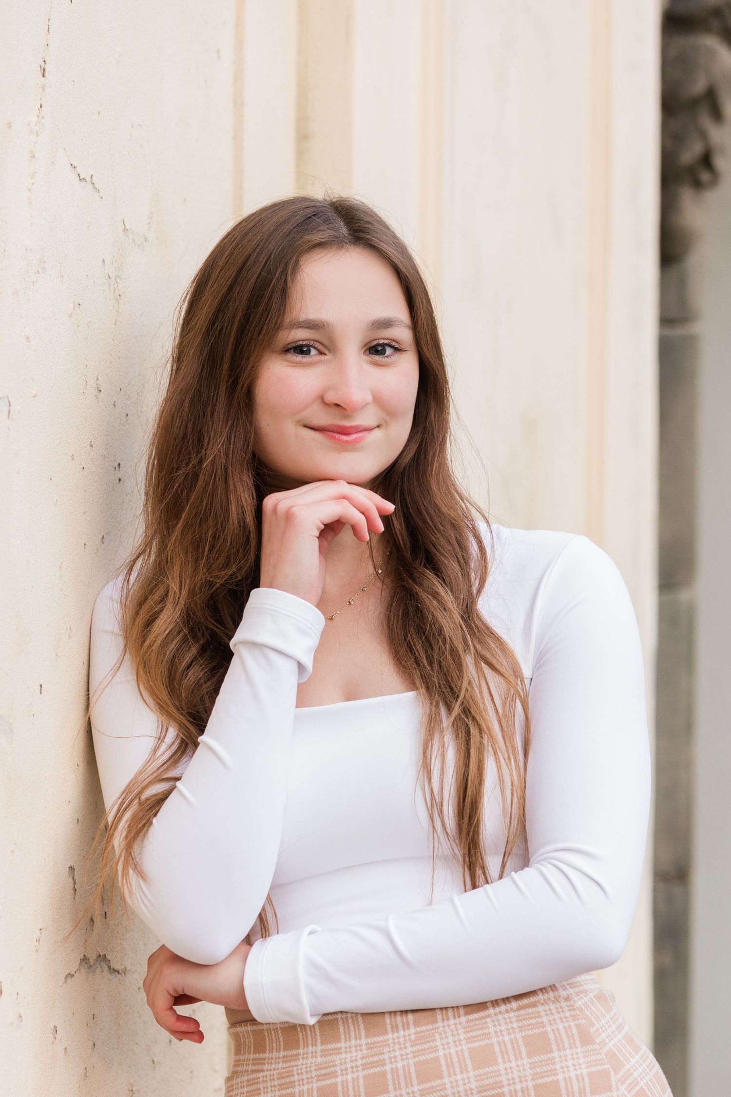 Young woman with long brown hair in white top smiling, resting chin on hand against a light stone wall.