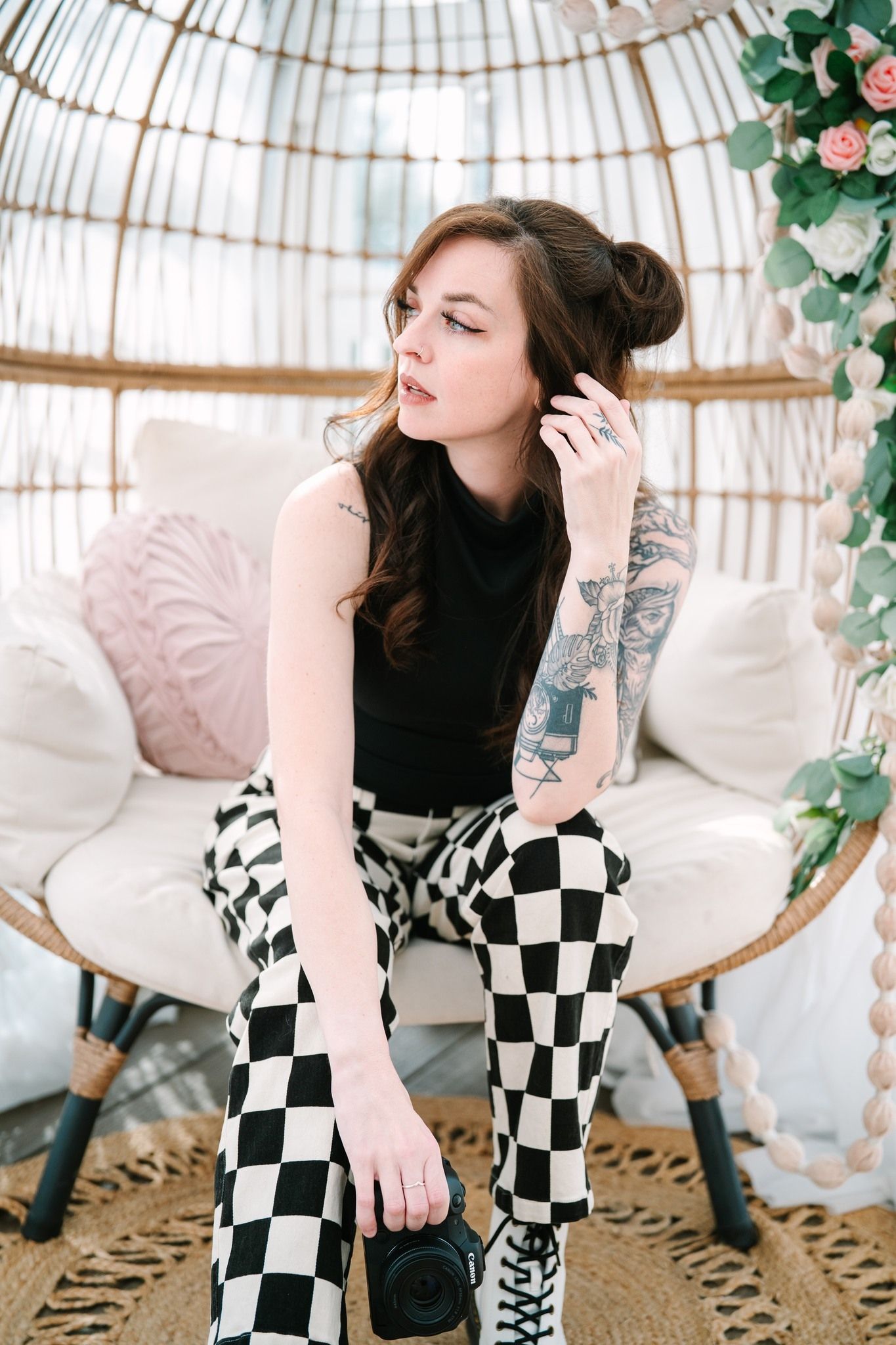 Young woman with tattoos in black top and checkered pants sits in wicker egg chair with floral decor.