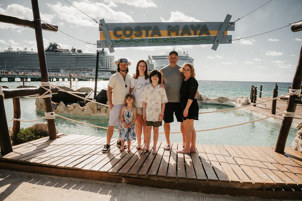 Family of six posing at Costa Maya cruise port sign with large ship and turquoise water in background.