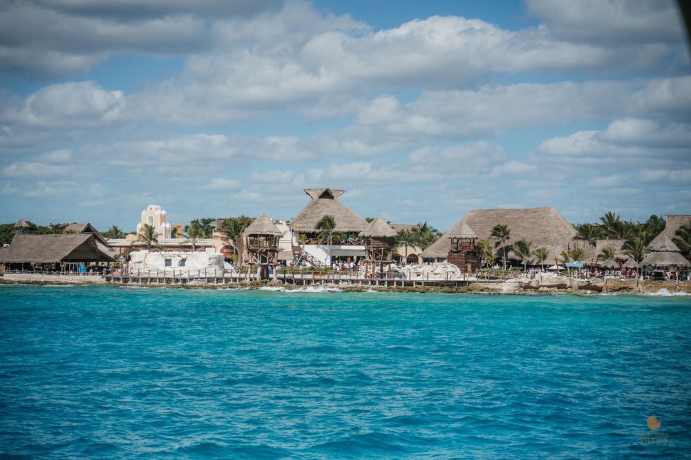 Turquoise Caribbean waters with thatched-roof resort buildings along a tropical coastline under partly cloudy skies.
