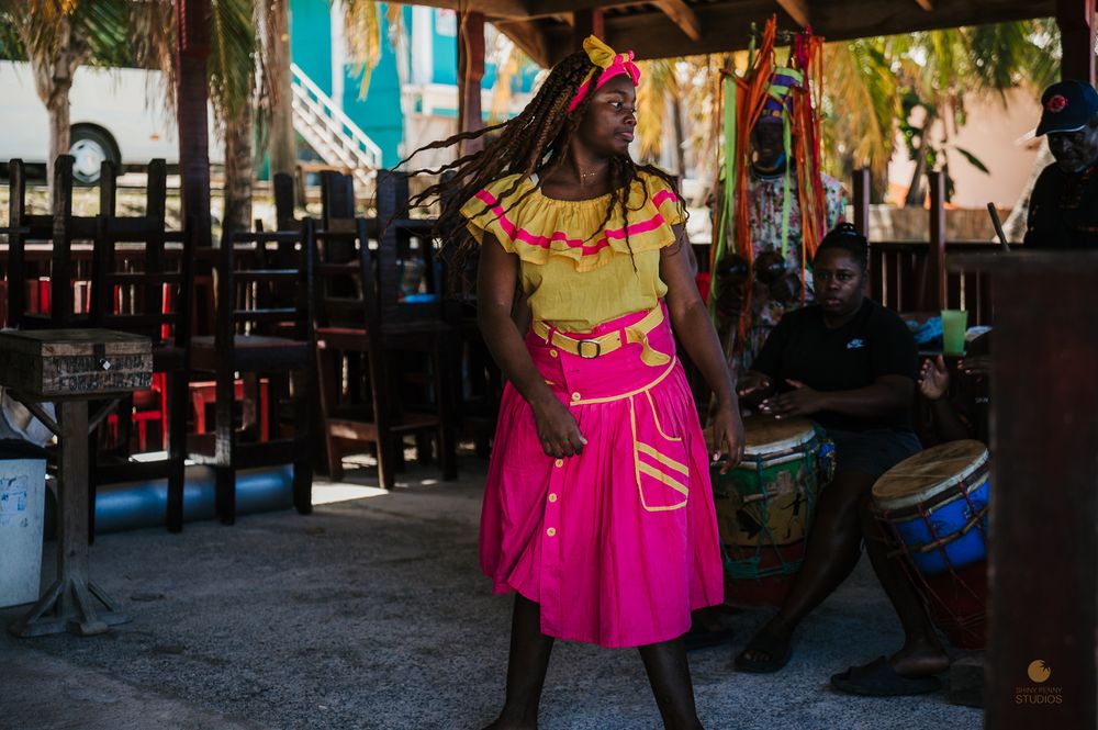 Colorful Caribbean dancer in bright pink skirt performs while drummer plays bongo drums outdoors.