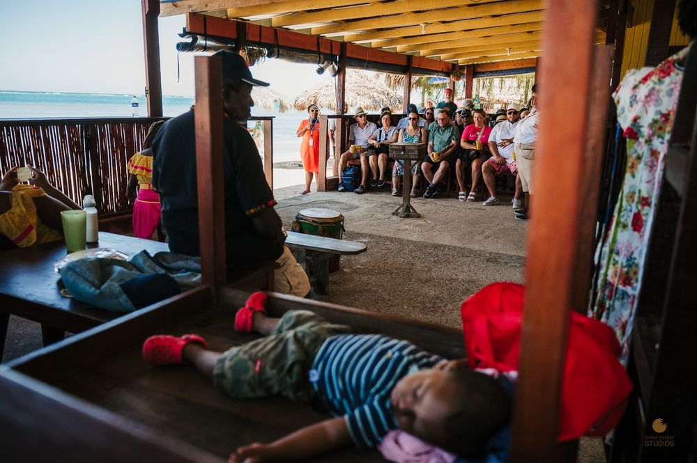 Child sleeping on a wooden bench while tourists sit in an open-air thatched pavilion by the beach.