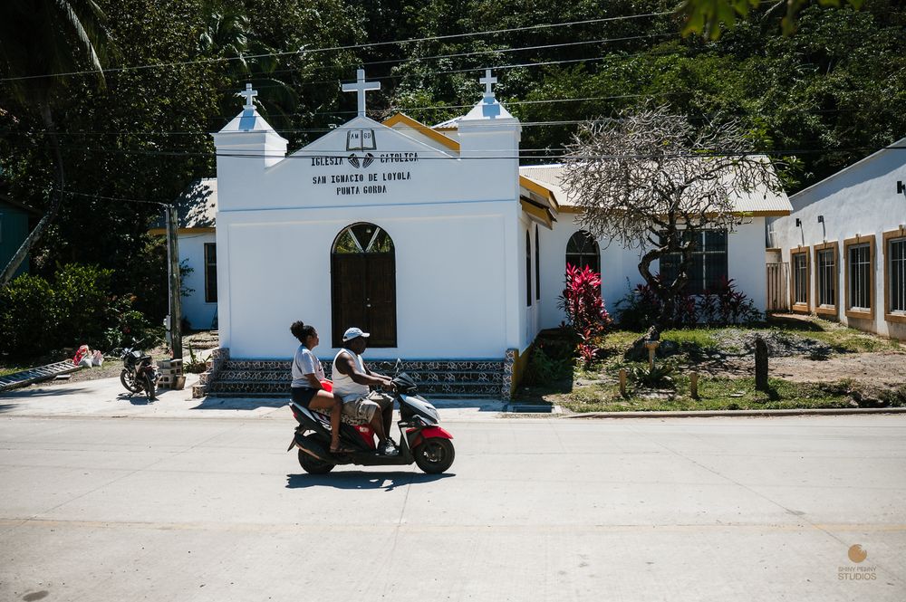 A red scooter passes a small white Catholic church on a quiet street in a tropical village.