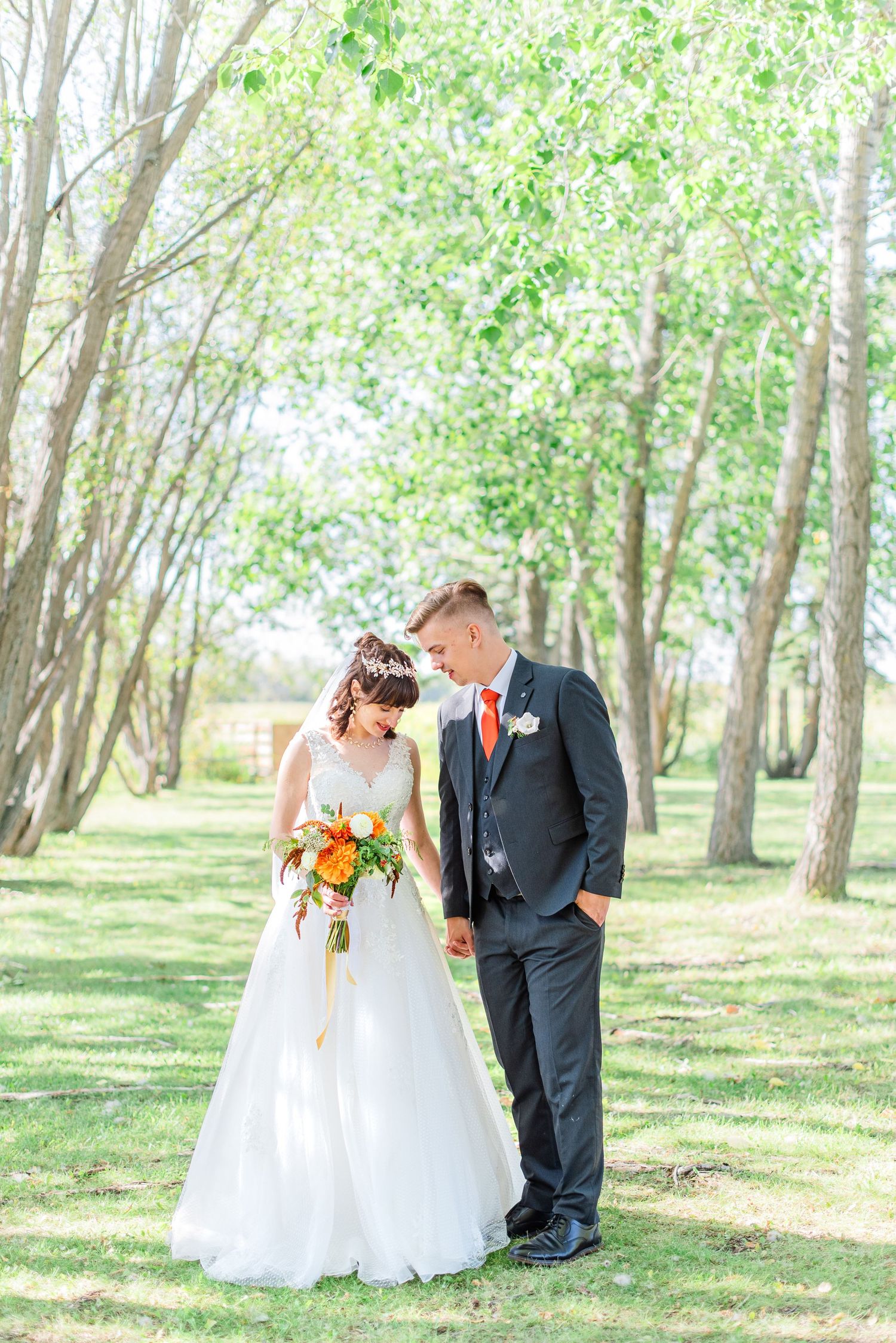 Bride and groom stand together in a sunlit grove, she holds an orange bouquet, wearing a white gown and floral crown.