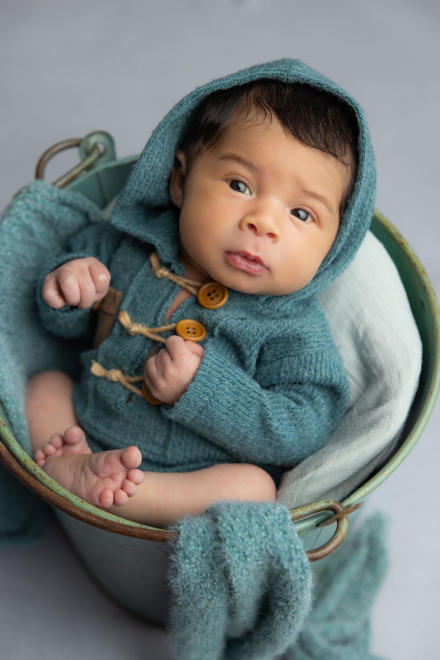 Newborn baby in teal hooded knit onesie with wooden buttons, sitting in a vintage metal bucket with soft teal blankets.