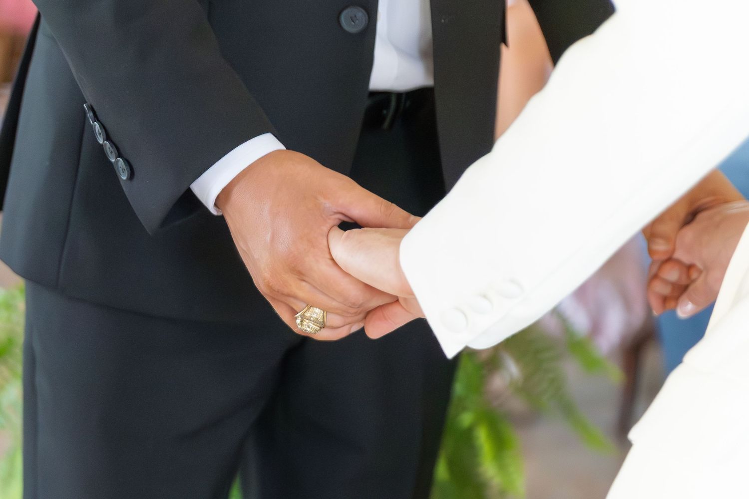 Couple holding hands during wedding ceremony, groom wearing gold ring and dark suit.