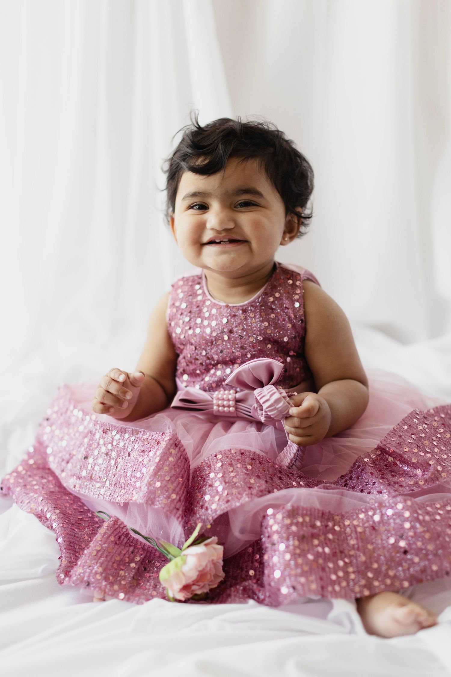 Smiling baby girl in a pink sequined dress with bow, sitting on white fabric, holding a pink flower.