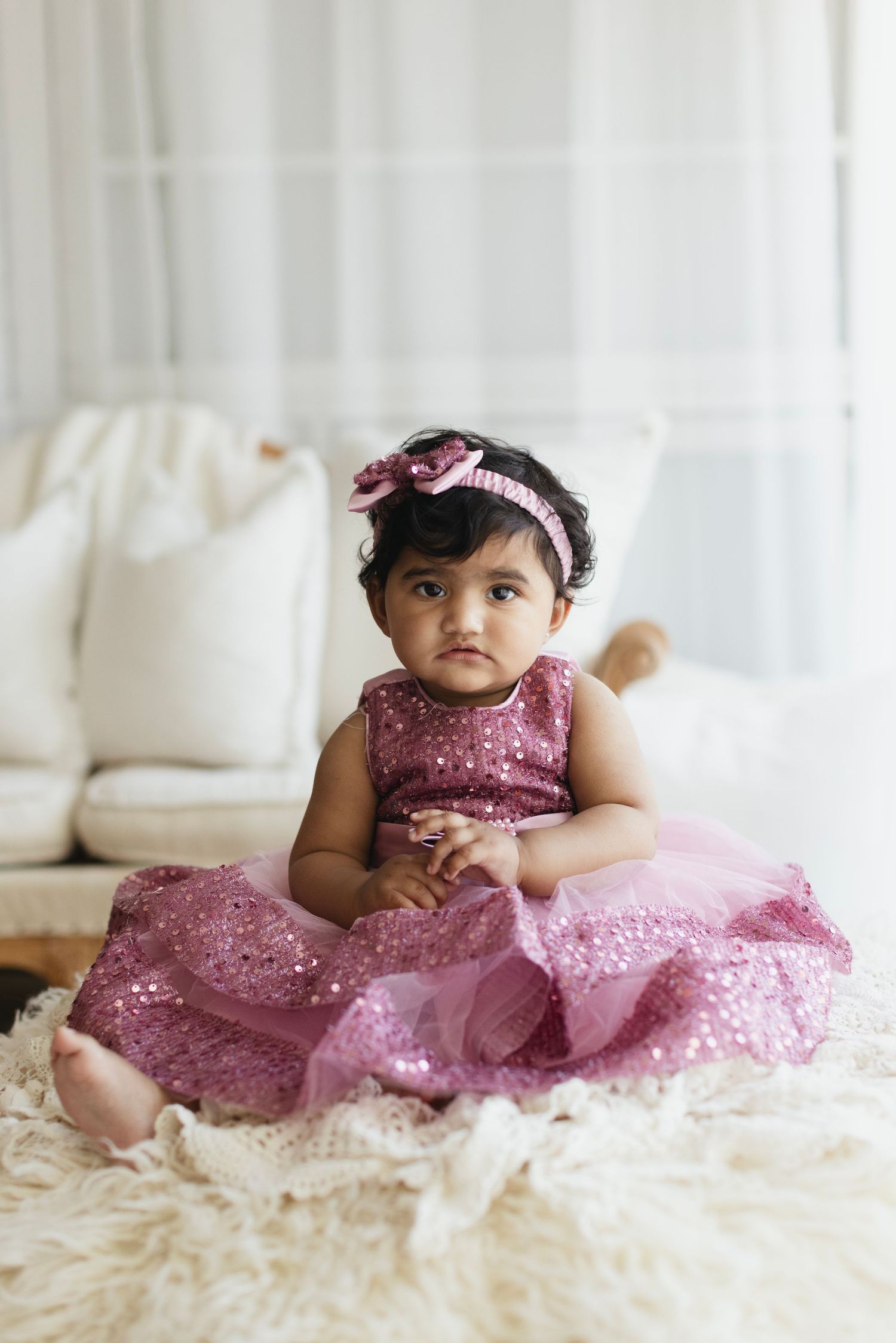 A baby girl in a sparkly pink dress and bow headband sits on a fluffy rug indoors.