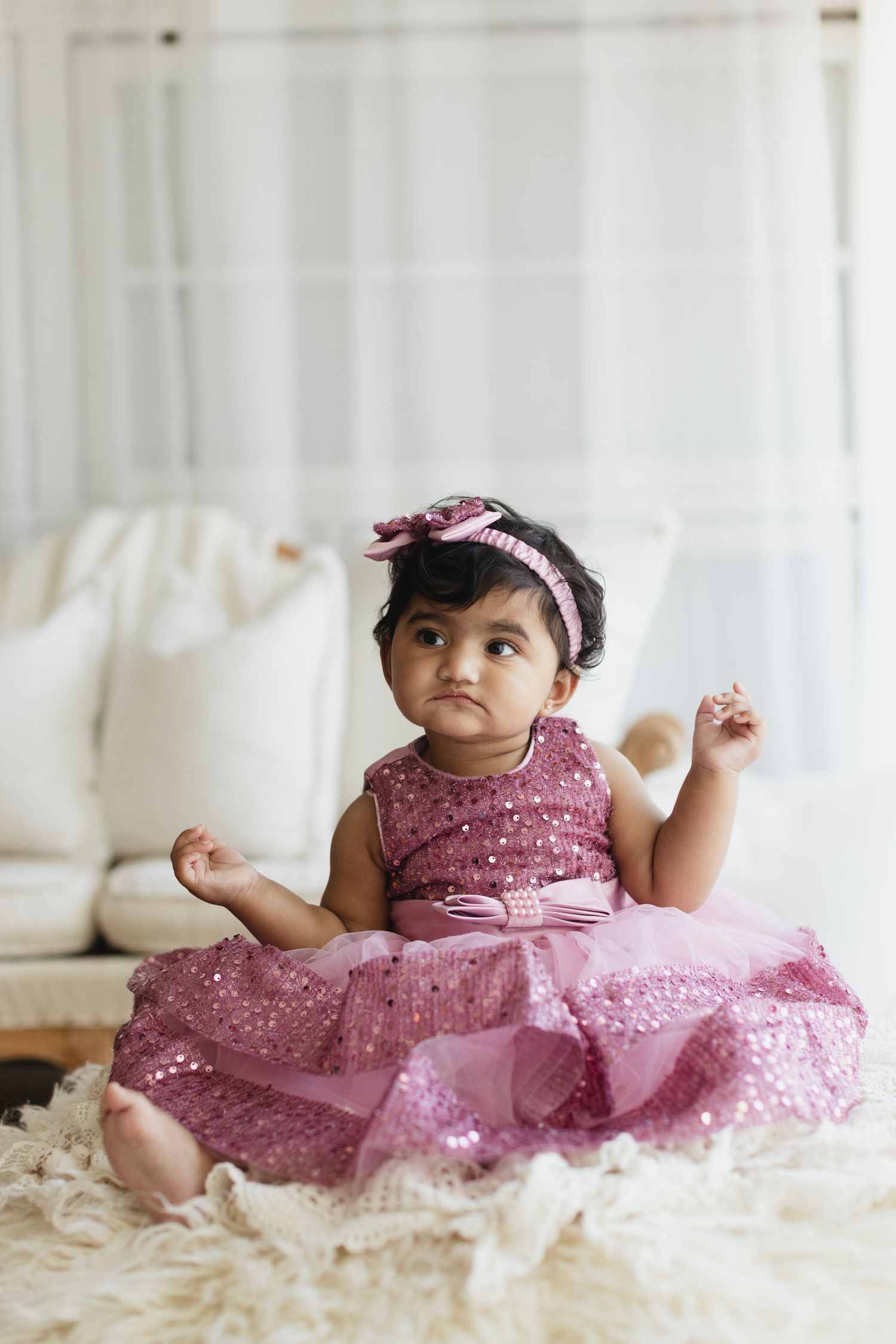 A cute baby girl in a pink sequin dress and bow headband sits on a fluffy rug in a bright, elegant room.